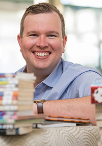 photograph of a mna from the shoulders up behind looking towards the camera from behind a stack of books.